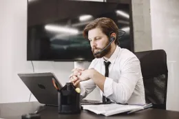 A bearded man wearing a black wireless headset with a boom microphone works at a desk with a laptop in an office.
