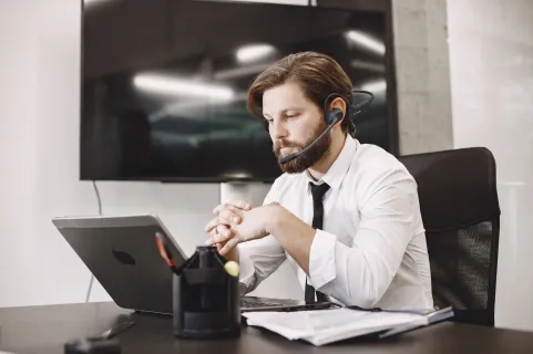 A bearded man wearing a black wireless headset with a boom microphone works at a desk with a laptop in an office.