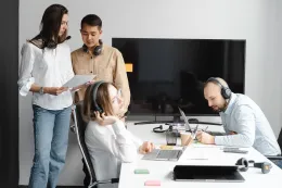 A group of customer support colleagues wearing headsets while discussing documents and working on laptops around an office desk.