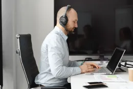 Side view of a man wearing an office headset with a boom microphone while working on a laptop at a desk.