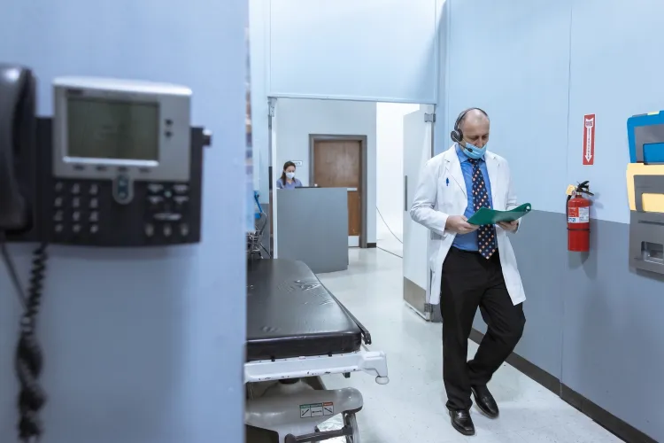 Male doctor wearing a white coat, face mask, and headset reading medical charts while walking down a clinic hallway.