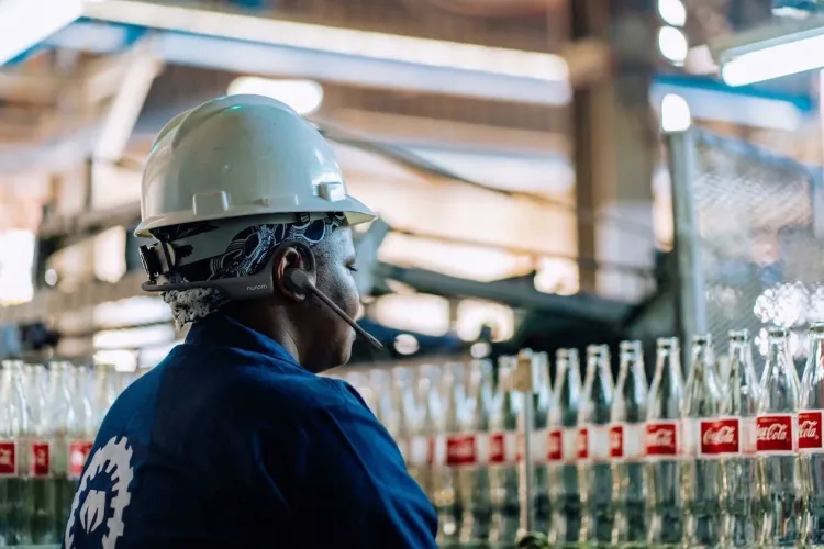 A worker in a manufacturing plant wears a hard hat and a Nuroum open-ear headset while standing in front of a bottle production line.