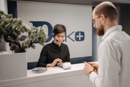 A female receptionist wearing a single-ear headset works at a front desk while assisting a male customer.
