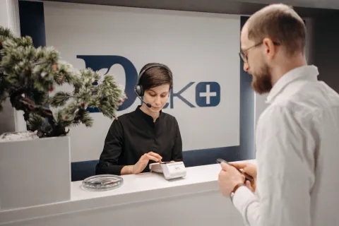 A female receptionist wearing a single-ear headset works at a front desk while assisting a male customer.