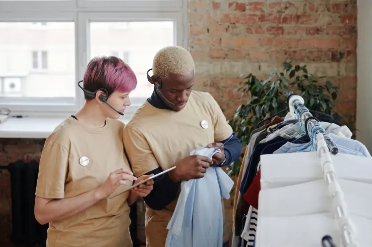 Two retail staff members wearing lightweight open-ear headsets with boom mics while managing clothing stock and using a tablet.