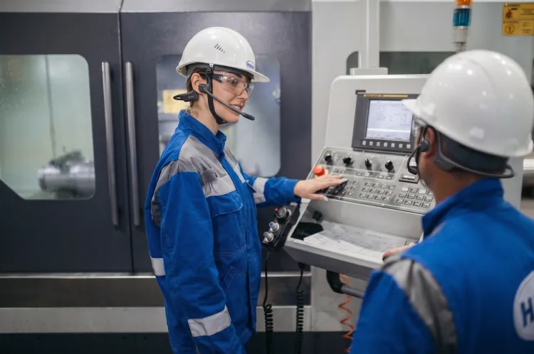 Two industrial technicians wearing hard hats and wireless boom mic headsets while operating a CNC machine control panel.