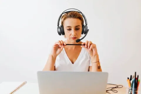 A woman in a white V-neck shirt, wearing a prominent black over-ear headset, focuses on her laptop while holding a small black stylus.