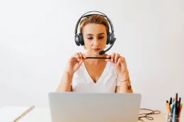 A woman in a white V-neck shirt, wearing a prominent black over-ear headset, focuses on her laptop while holding a small black stylus.